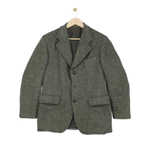 Pode incluir: Um blazer de manga comprida com um padrão de espinha de peixe em verde azeitona escuro e castanho. A peça apresenta uma lapela com entalhe, três botões castanhos escuros na frente, dois bolsos com aba na cintura e um bolso no peito, pendurado num cabide de madeira.