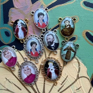 Puede incluir: Una colección de colgantes ovalados con figuras religiosas. Los colgantes tienen marcos dorados y plateados, con imágenes de santos y otras figuras religiosas. El fondo es una mezcla de verde azulado, dorado y amarillo.
