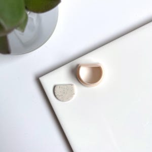 Peut inclure: Deux boucles d'oreilles en céramique en forme de demi-cercle, l'une avec un glaçage blanc moucheté et l'autre avec un glaçage rose clair uni.