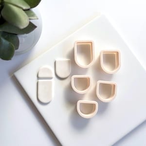 Op de afbeelding: Een verzameling beige polymeerklei-uitsteekvormen in verschillende vormen, waaronder afgeronde rechthoeken en een pilvorm, gerangschikt op een wit oppervlak. Een kleine groene plant in een witte pot is zichtbaar.