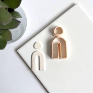 Op de afbeelding: Twee koekjesvormen in de vorm van de letter "i" met afgeronde toppen. De vormen zijn gemaakt van lichtroze plastic.