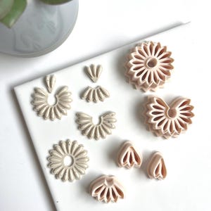 Op de afbeelding: Een verzameling beige polymeerklei oorbelcomponenten in verschillende bloem- en waaier vormen. De componenten zijn gerangschikt op een wit oppervlak en tonen een verscheidenheid aan ontwerpen, waaronder cirkelvormige, halve cirkel en bloembladachtige vormen. De klei heeft een gespikkelde textuur.