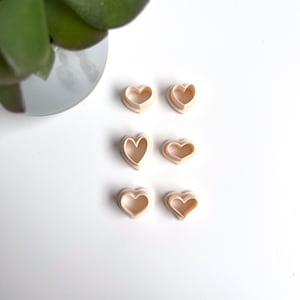 Könnte beinhalten: Sechs herzförmige Ausstecher aus hellbeigefarbenem Kunststoff sind in drei Reihen zu zweit auf einer weißen Oberfläche angeordnet.