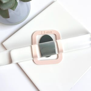 Peut inclure: Un rouleau à pâtisserie en acrylique transparent avec un cadre rose marqué "2,5 mm" est centré sur une forme ovale gris foncé. Le rouleau est posé sur une surface blanche, avec une petite plante verte dans un pot gris en arrière-plan.