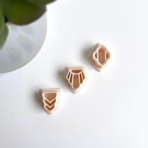 Peut inclure: Trois emportes-pièces en plastique rose de formes géométriques différentes. Les formes sont un diamant, une larme et un chevron.