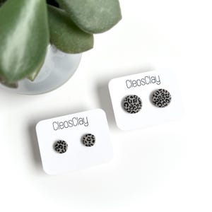 Könnte beinhalten: Zwei Paar runde Ohrstecker mit Leopardenmuster auf weißen Karten. Die Karten sind mit "CleosClay" beschriftet. Die Ohrringe sind grau mit schwarzen Punkten. Im Hintergrund ist eine grüne Pflanze zu sehen.