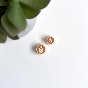 Op de afbeelding: Een paar kleine, ronde, bruine en witte oorbellen met een gestileerd tarwemotief.