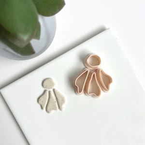 Op de afbeelding: Twee beige oorbeluitstekers van polymeerklei in een bloemmotief. Eén uitsteker is een afgewerkte oorbel, de andere is een uitsteker. De uitstekers liggen op een wit oppervlak met een plant op de achtergrond.