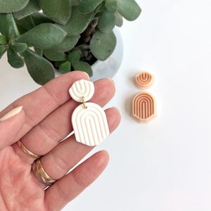 May include: White and peach polymer clay earrings with a geometric arch design. The earrings are dangle style and have a gold hook.