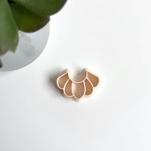 Peut inclure: Un emporte-pièce de couleur pêche clair en forme de fleur stylisée ou de pétales. Le découpoir est en plastique et repose sur une surface blanche. Le motif présente un dessus incurvé avec cinq formes de pétales arrondies.