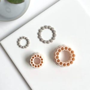 Könnte beinhalten: Vier kreisförmige Ausstecher in verschiedenen Größen. Zwei sind hellgrau und zwei sind hellpfirsichfarben. Jeder Ausstecher hat einen Perlenrand, der ein dekoratives Design erzeugt. Die Ausstecher sind auf einer weißen Oberfläche angeordnet.