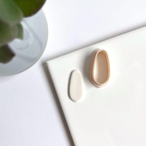 Op de afbeelding: Twee witte kleivormige vormen, een eenvoudige ovaal en de andere een meer organische, vrije vorm. De vormen bevinden zich op een wit oppervlak.