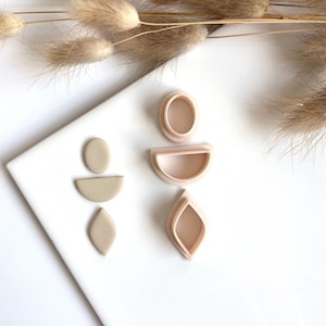 Op de afbeelding: Een verzameling polymeerklei oorbelvormen in verschillende geometrische vormen, waaronder een ovaal, een halve cirkel en een druppel. De vormen zijn lichtroze en de kleistukken zijn beige. Gedroogd siergras op de achtergrond.