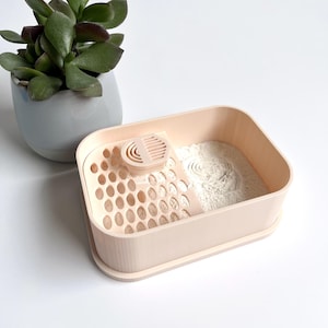 Op de afbeelding: Een lichtroze plastic container met een uitneembaar dienblad en een kleine schep. Het dienblad heeft een rooster met gaten en de container is gevuld met wit poeder.