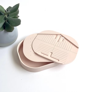 Peut inclure: Un organisateur de bijoux rose clair avec un couvercle et plusieurs compartiments. Le couvercle présente un motif géométrique avec des lignes et des découpes. L'organisateur est sur une surface blanche, avec une petite plante en pot en arrière-plan.