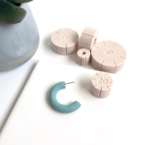 Op de afbeelding: Een turquoisekleurige oorring met een zilveren pin ligt naast verschillende beige, cilindrische gereedschappen voor het maken van sieraden. De gereedschappen zijn gemarkeerd met nummers, waaronder 20, 35 en 40. De oorring en gereedschappen liggen op een wit oppervlak.