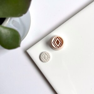 Peut inclure: Deux petits carreaux de céramique avec des motifs géométriques. Un carreau est blanc avec un motif de diamant gris, et l'autre est pêche avec un motif de diamant concentrique.