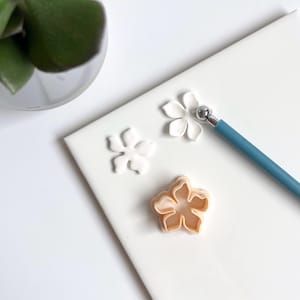 Op de afbeelding: Een close-up van een witte keramische tegel met twee witte fondantbloemen en een bloemvormige koekjesuitsteker. Een blauw gereedschap met een bolvormige punt is ook zichtbaar.