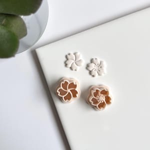 Op de afbeelding: Drie plastic koekjesvormen in de vorm van kersenbloesems. De vormen zijn in verschillende maten en zijn lichtroze van kleur.