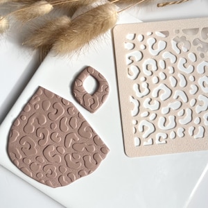Op de afbeelding: Een verzameling knutselspullen met een luipaardprint sjabloon in beige, een druppelvormige oorbel en een groot, onregelmatig gevormd stuk, allemaal in een gedempte bruine kleur. De oorbel heeft een gat in het midden. De stukken hebben een textuur luipaardpatroon.