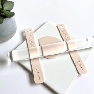 Könnte beinhalten: Eine transparente Acryl-Tonrolle mit zwei beigefarbenen 2-mm-Abstandshaltern und einem beigefarbenen Tonkreis auf einer weißen Fliese. Die Abstandshalter sind mit "CleosClay" beschriftet. Eine kleine Sukkulentenpflanze in einem grauen Topf befindet sich im Hintergrund.