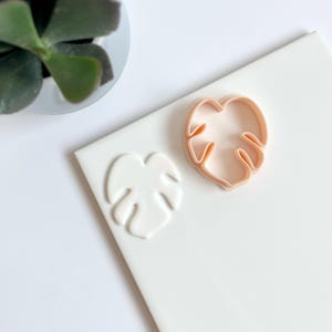 Peut inclure: Gros plan sur deux emporte-pièces en forme de feuilles de monstera. Les emporte-pièces sont en plastique rose clair et se trouvent sur une surface blanche.