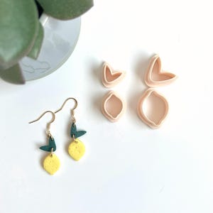 Peut inclure: Boucles d'oreilles en forme de citron avec des crochets dorés, des citrons jaunes et des feuilles vertes. Quatre emporte-pièces en argile rose clair en forme de citron sont également présents. Accessoire unique.