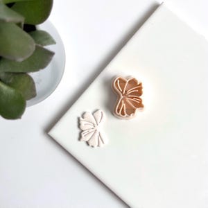 Op de afbeelding: Twee plastic koekjesvormen in de vorm van een strik. De vormen zijn lichtroze en liggen op een wit oppervlak.