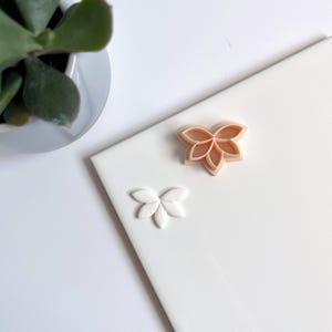 Op de afbeelding: Twee plastic koekjesvormen in de vorm van een bloem. De bovenste vorm is perzikkleurig en de onderste vorm is wit.