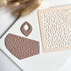 Könnte beinhalten: Bastelmaterialien für Ohrringe und Accessoires. Das Bild zeigt einen tropfenförmigen Ohrring, ein rechteckiges Stück und eine Schablone, alle in einem gedämpften Braunton mit Leopardenmuster. Die Artikel sind auf einer weißen Oberfläche angeordnet.