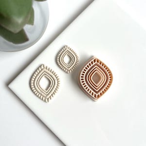 Op de afbeelding: Drie beige kleisnijders in een oog- of blad vorm. De snijders hebben een gelaagd ontwerp met een getextureerde rand. De grootste snijder heeft meerdere lagen, terwijl de kleinere er één hebben. De snijders liggen op een wit oppervlak.