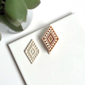 Peut inclure: Deux emporte-pièces en forme de losange avec des motifs géométriques. L'un est de couleur beige clair et l'autre est beige plus foncé. Les emporte-pièces sont placés sur une surface blanche à côté d'une plante verte.