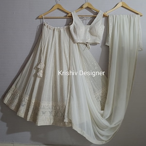 May include: A white, sequined and embroidered lehenga with a matching white, sequined and embroidered crop top. The outfit is paired with a white chiffon dupatta. The text "Krishiv Designer" is visible on the skirt.