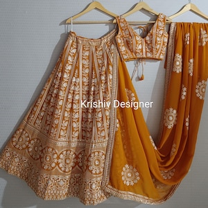 May include: A mustard yellow embroidered lehenga with a matching embroidered blouse and dupatta. The lehenga has a flared skirt with a white embroidered design. The blouse has a V-neckline and a tie-up closure. The dupatta is a sheer fabric with a matching embroidered design. The text "Krishiv Designer" is visible on the image.
