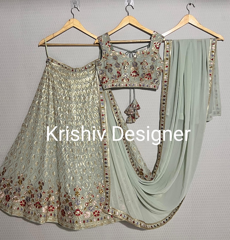 May include: A light green three-piece Indian bridal outfit with gold embroidery and red and silver embellishments. The outfit includes a long skirt, a sleeveless blouse, and a matching dupatta.