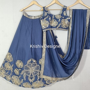 May include: A three-piece ensemble featuring a blue skirt, a matching sleeveless top, and a draped scarf. The skirt and top are adorned with gold floral embroidery and a gold border. The text "Krishiv Designer" is visible.