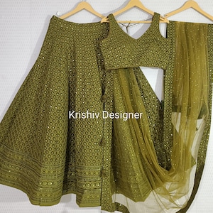 May include: Olive green three-piece ensemble featuring a skirt, a sleeveless top, and a sheer scarf. The skirt and top are adorned with a diamond pattern and small embellishments. The text "Krishiv Designer" is visible.