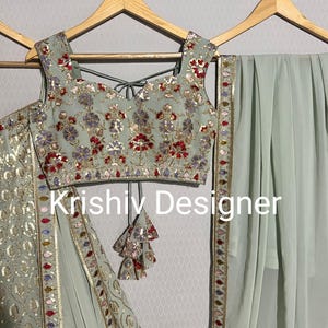 May include: A sage green embroidered crop top with a matching sage green skirt and dupatta. The top has a V-neckline and is decorated with silver sequins, beads, and floral embroidery. The skirt has a similar design and is paired with a matching dupatta. The dupatta has a border of silver sequins and beads. The text "Krishiv Designer" is visible on the image.