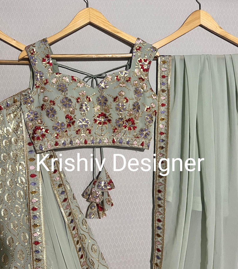 May include: A sage green embroidered crop top with a matching sage green skirt and dupatta. The top has a V-neckline and is decorated with silver sequins, beads, and floral embroidery. The skirt has a similar design and is paired with a matching dupatta. The dupatta has a border of silver sequins and beads. The text "Krishiv Designer" is visible on the image.