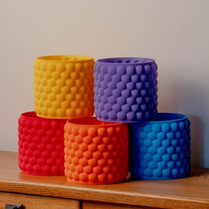 May include: Four colourful cylindrical planters with a textured bubble design. The planters are stacked in a pyramid shape, with the yellow planter on top, followed by the purple, then the orange, and finally the blue planter.