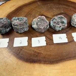 May include: Five rough-cut gemstones with a mix of pink and green hues are arranged on a wooden surface. Each stone is unique in shape and texture. Small white labels numbered 1 through 5 are placed below each stone.
