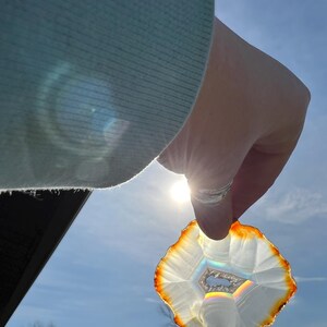 May include: A white agate slice with a rainbow iridescence held up to the sun. The agate has a brown outer edge and a white centre with a hexagonal pattern.