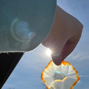 May include: A white agate slice with a rainbow iridescence held up to the sun. The agate has a brown edge and a natural, irregular shape.
