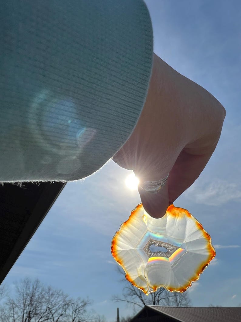 May include: A white agate slice with a rainbow iridescence held up to the sun. The agate has a brown edge and a natural, irregular shape.
