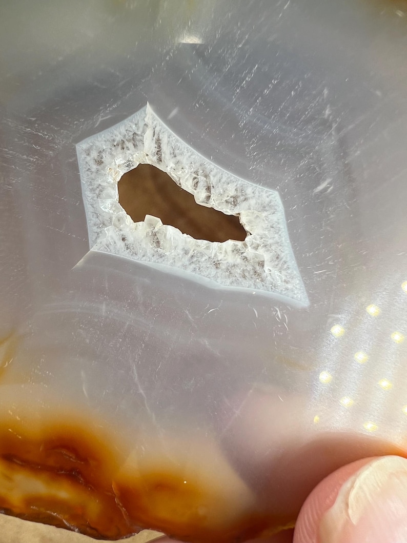 May include: Close-up of a light grey agate slice with a large, irregular hole in the centre. The agate has a brown band around the edge.