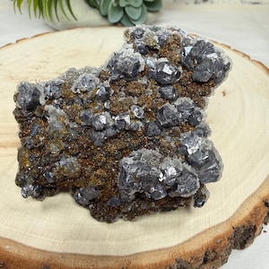 May include: A mineral specimen featuring metallic, dark gray cubic crystals clustered on a textured, bronze-colored base. The cluster displays a mix of sharp, reflective geometric shapes and granular, earthy surfaces, creating a complex, multi-dimensional geological formation.