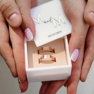 May include: A white ring box containing two gold wedding bands. The box lid features the text "M and M" and the date "08/06/2024". The rings are presented in a beige interior. The image is held by two hands.