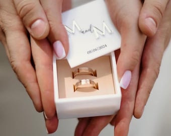 May include: A white ring box with two gold wedding bands. The box lid has the text "M and M" and the date "08/06/2024". The rings are displayed in a beige interior. The image is held by two hands.