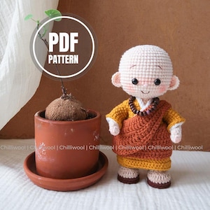Op de afbeelding: Gehaakte monnikpop in gele en oranje gewaden, met een kralenketting. De pop heeft een kale kop en een vredige uitdrukking. Links staat een kleine potplant en de tekst "PDF PATTERN" staat in een cirkel.