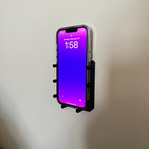 Può includere: Un supporto per telefono nero montato su una parete bianca. Il telefono mostra uno sfondo sfumato viola con l'ora 1:58 e la data domenica 25 febbraio.